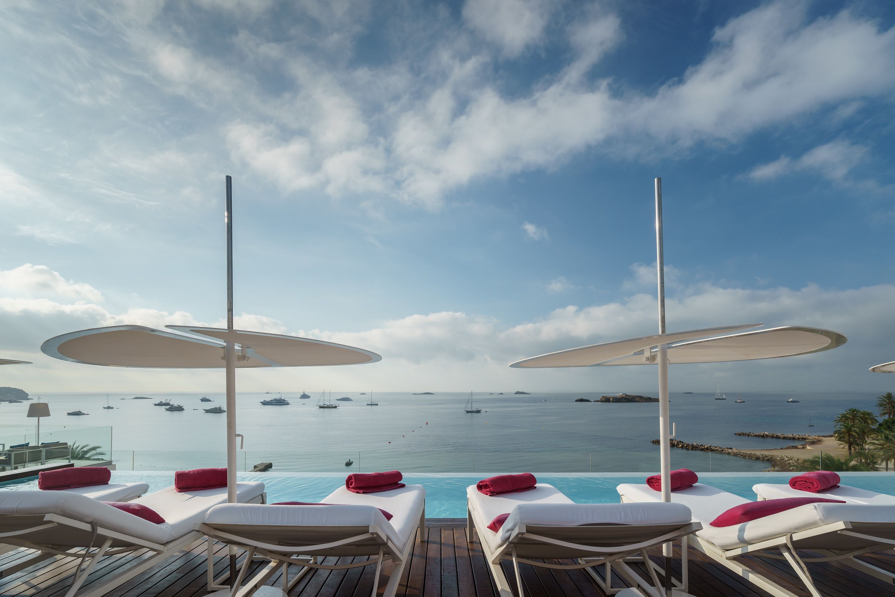 A poolside area with lounge chairs and umbrellas, overlooking a calm sea with boats.