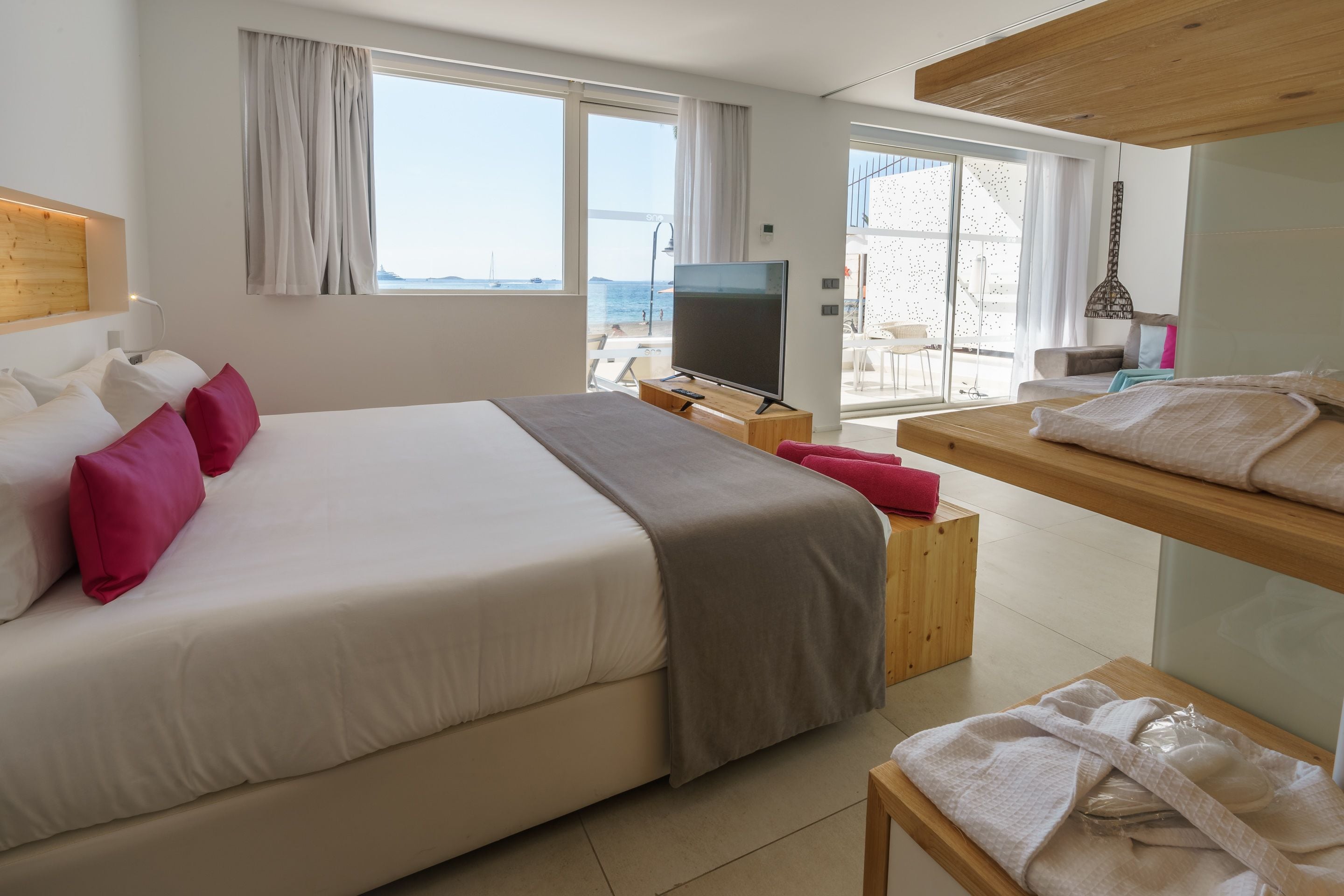 Habitación de hotel con una cama grande, televisión, banco de madera y balcón con vista al mar.