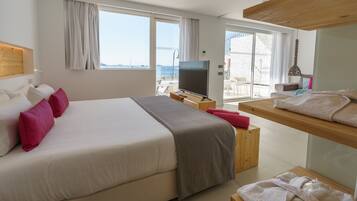 Habitación de hotel con una cama grande, televisión, banco de madera y balcón con vista al mar.