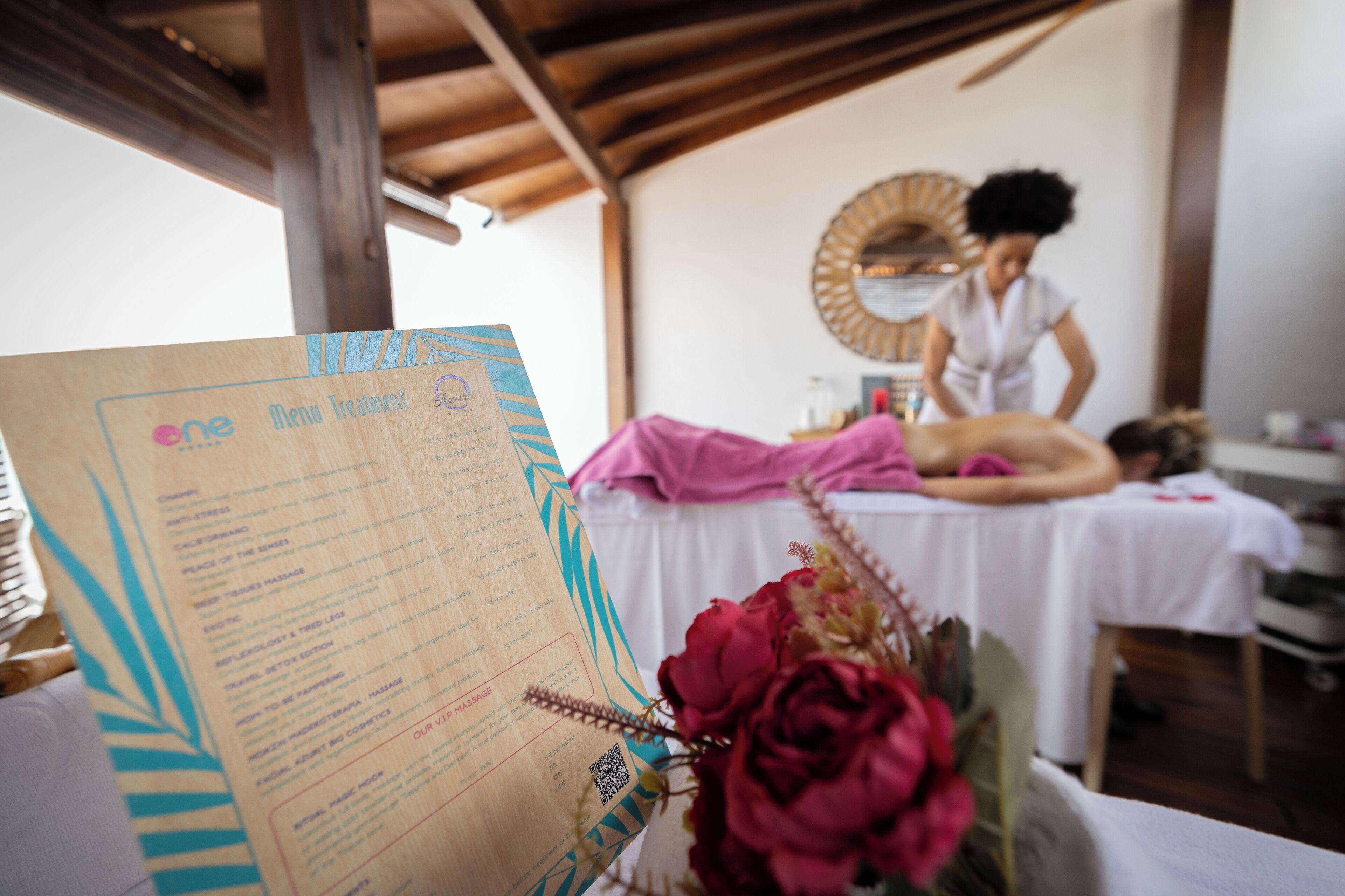Une salle de massage équipée d’une table de massage en bois, d’une autre table de massage recouverte d’une serviette rose, et d’un présentoir avec une carte des soins.