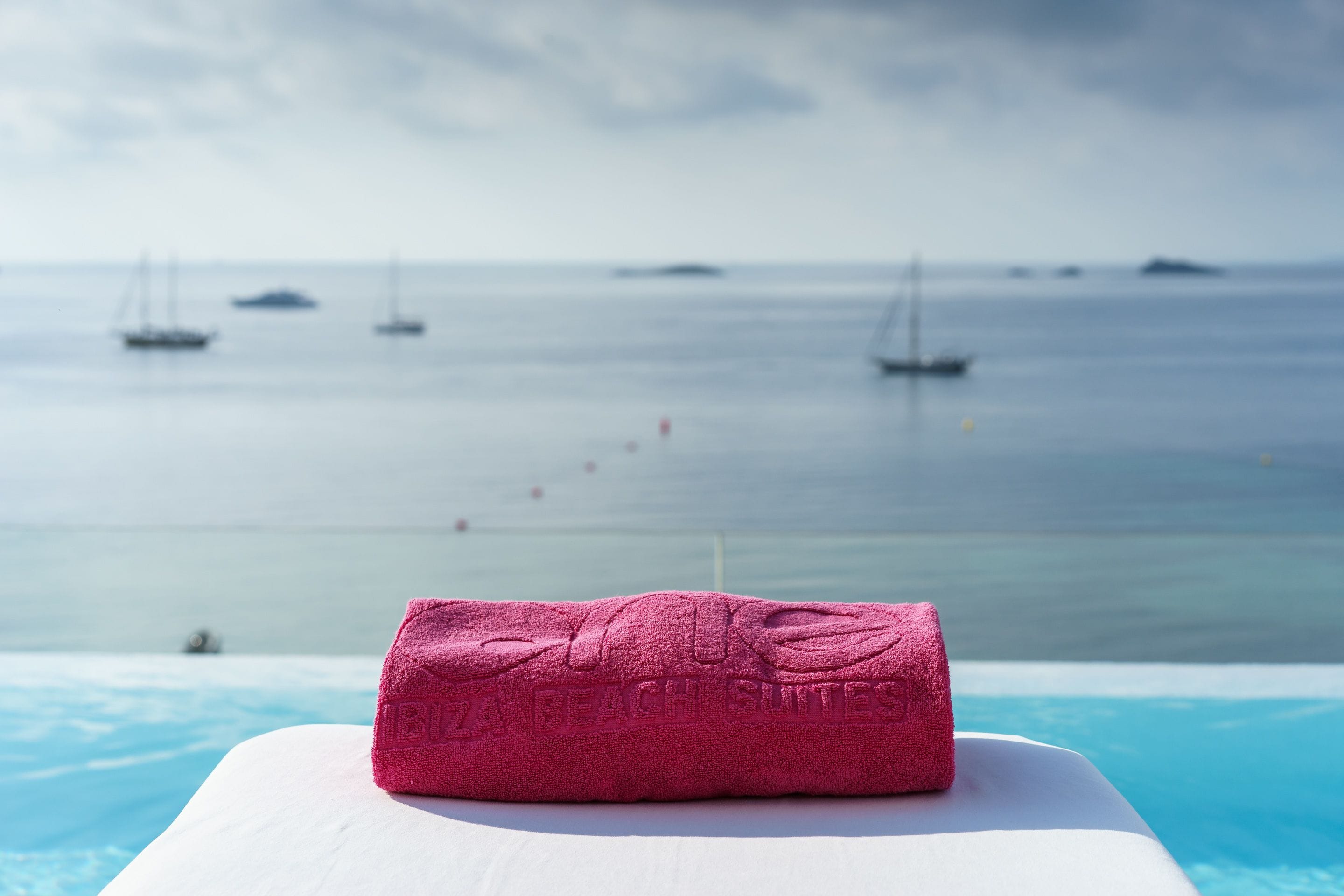 A pink towel on a white lounge chair by a pool with a view of the sea and boats.