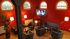 A modern living room with red walls, wooden flooring, and a flat-screen TV mounted on the wall. There are several black sofas and a coffee table, along with a potted plant and two floor lamps.