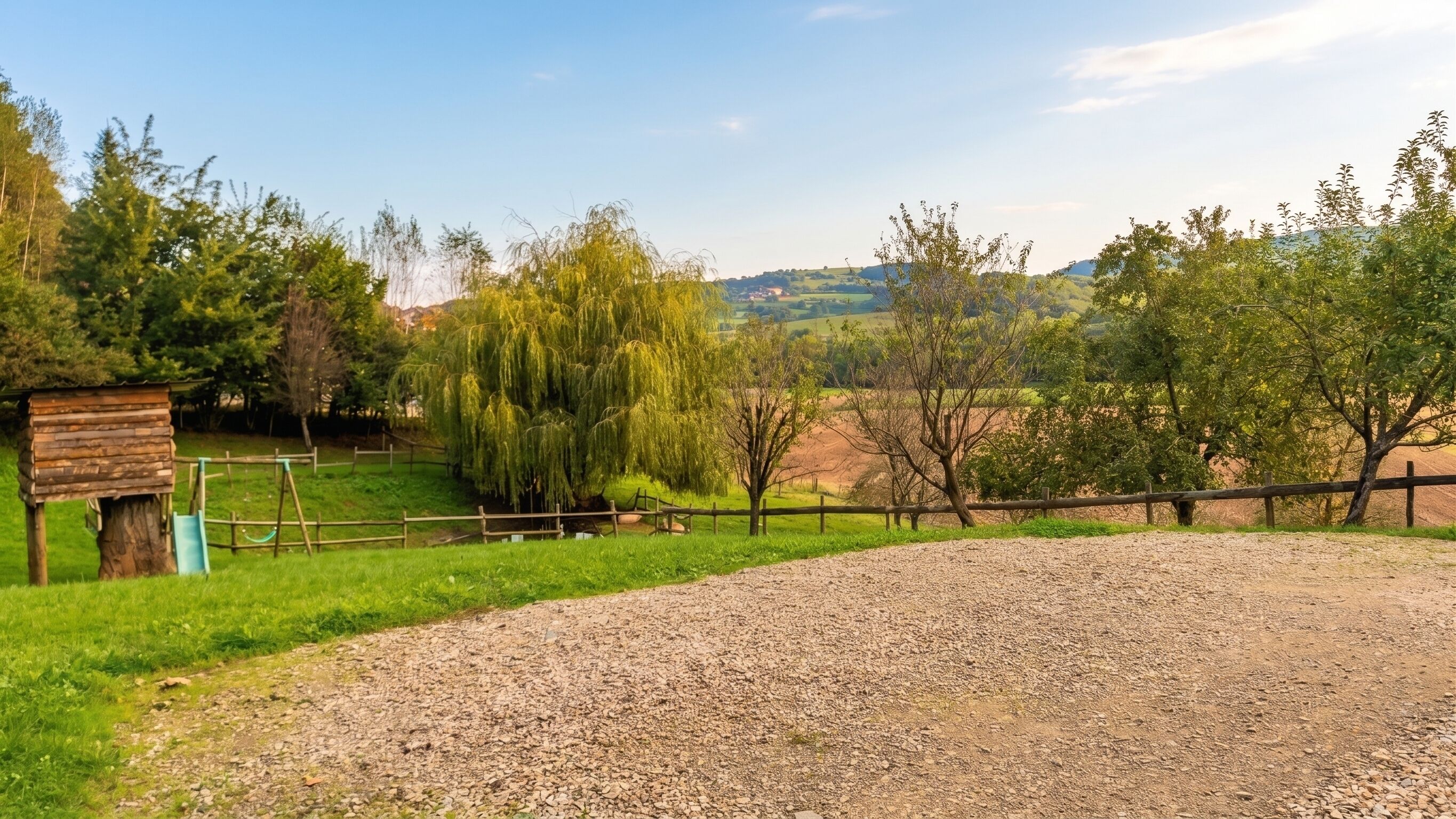 A gravel path leading to a wooden fence, a wooden structure, and a weeping willow tree.