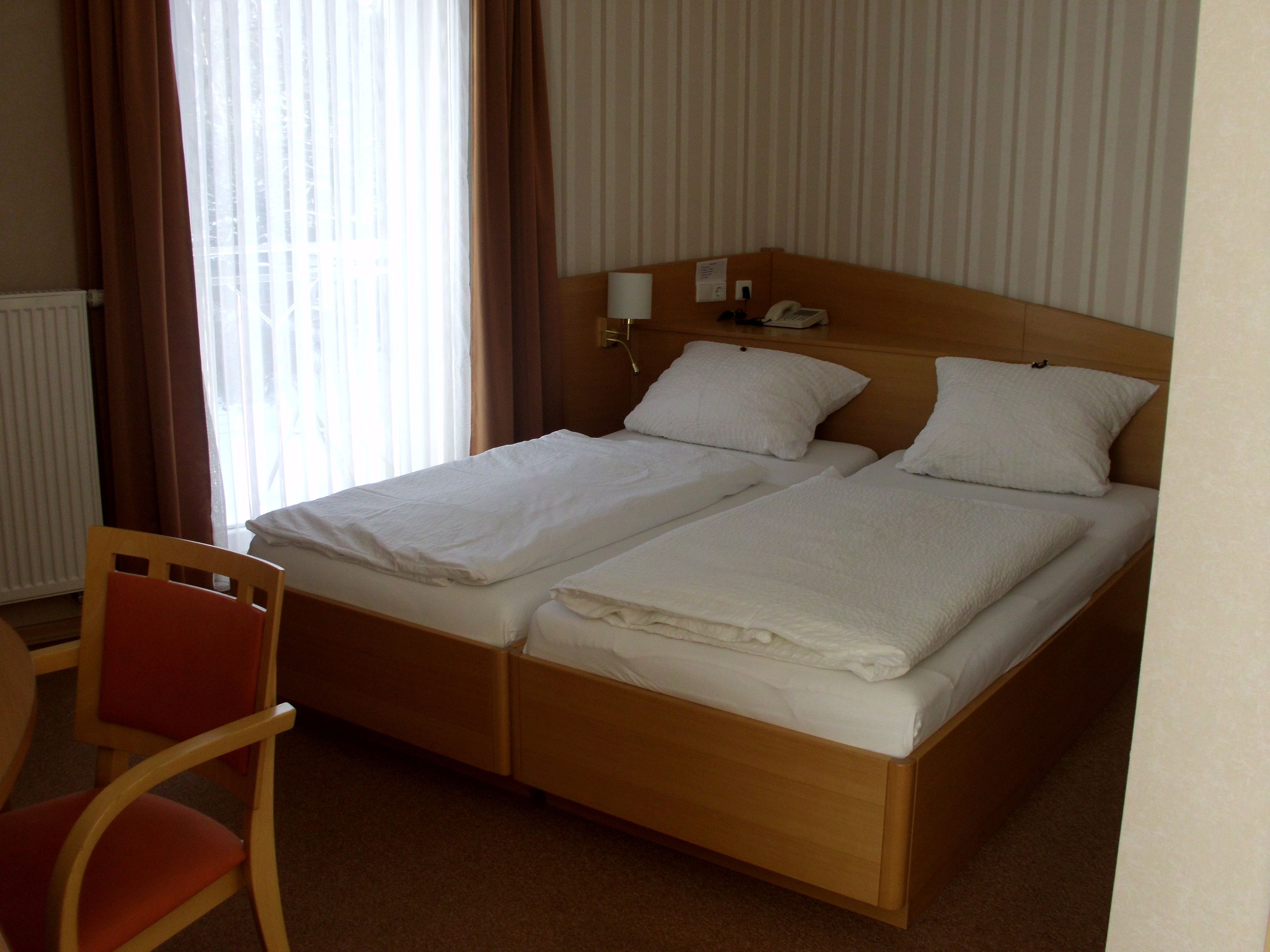 Cama doble con cabecera de madera, dos almohadas blancas, una mesita de noche con lámpara y una silla.