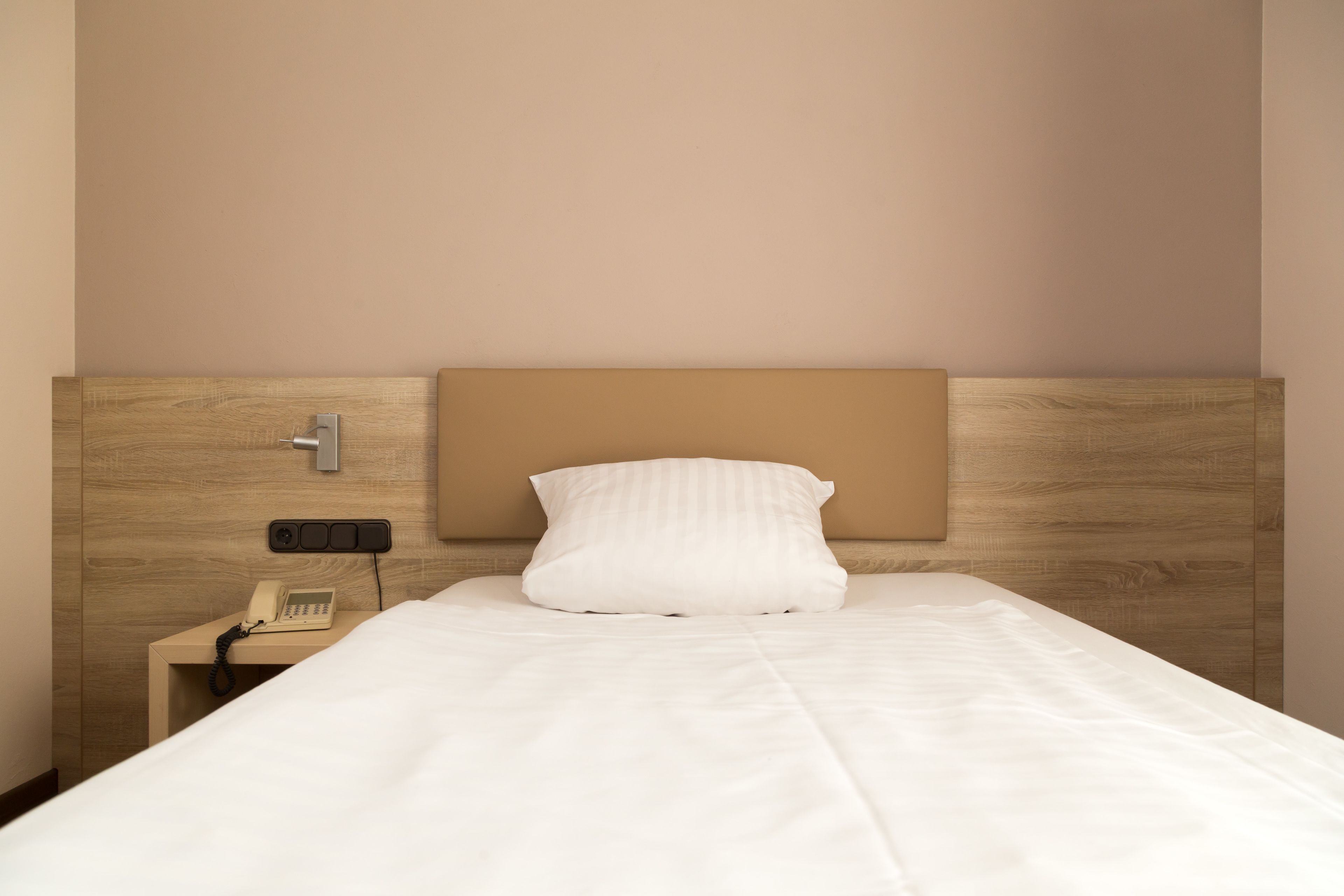A neatly made bed with a headboard, a bedside telephone, and a wall-mounted light switch.