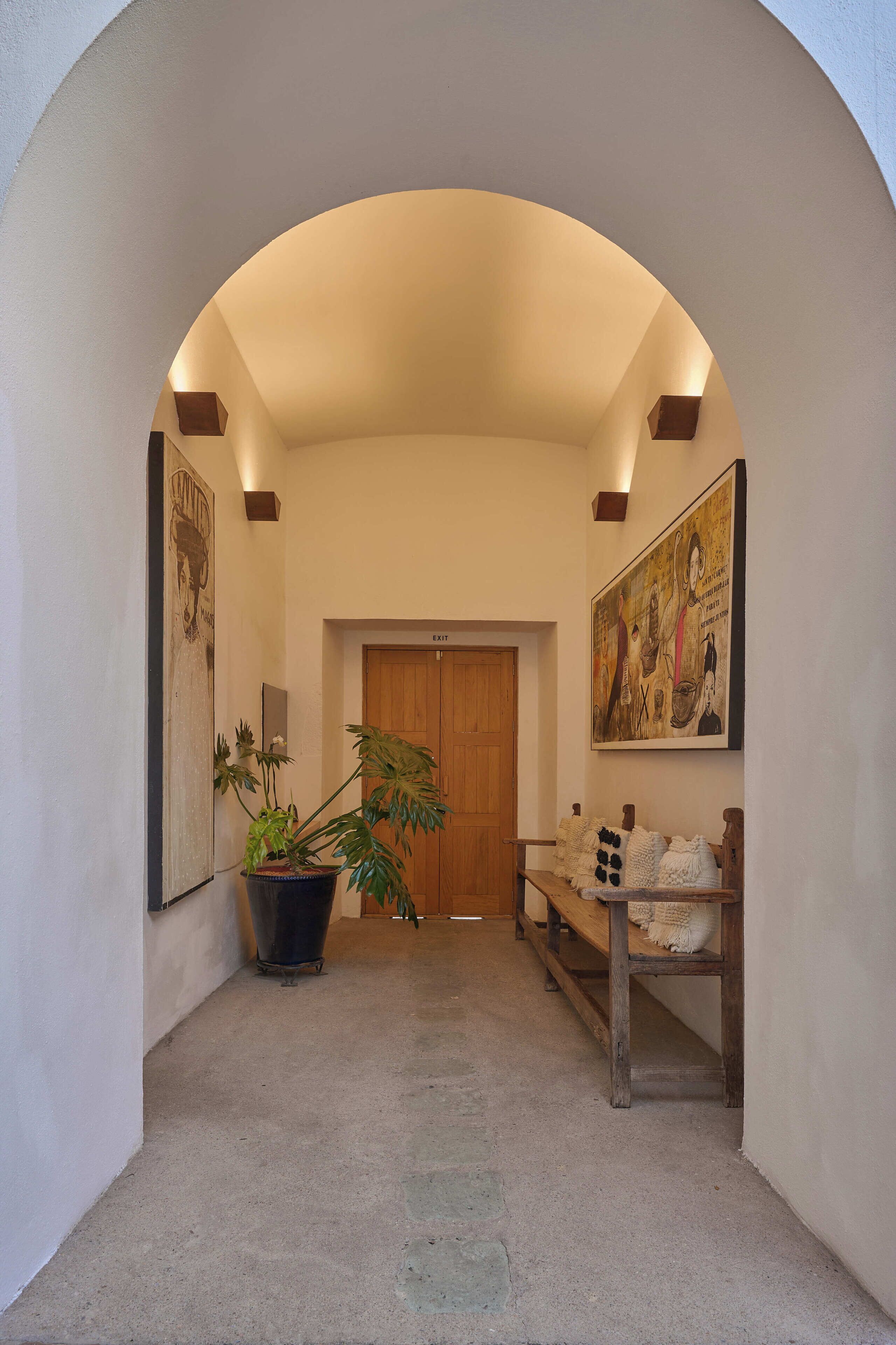 A hallway with a wooden door, a large potted plant, a bench with cushions, and a framed picture on the wall.