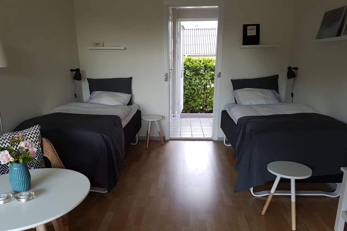 Ein Zimmer mit zwei Betten, einem kleinen Tisch und einem Stuhl. Eine Tür führt zu einem Außenbereich mit Grünpflanzen.