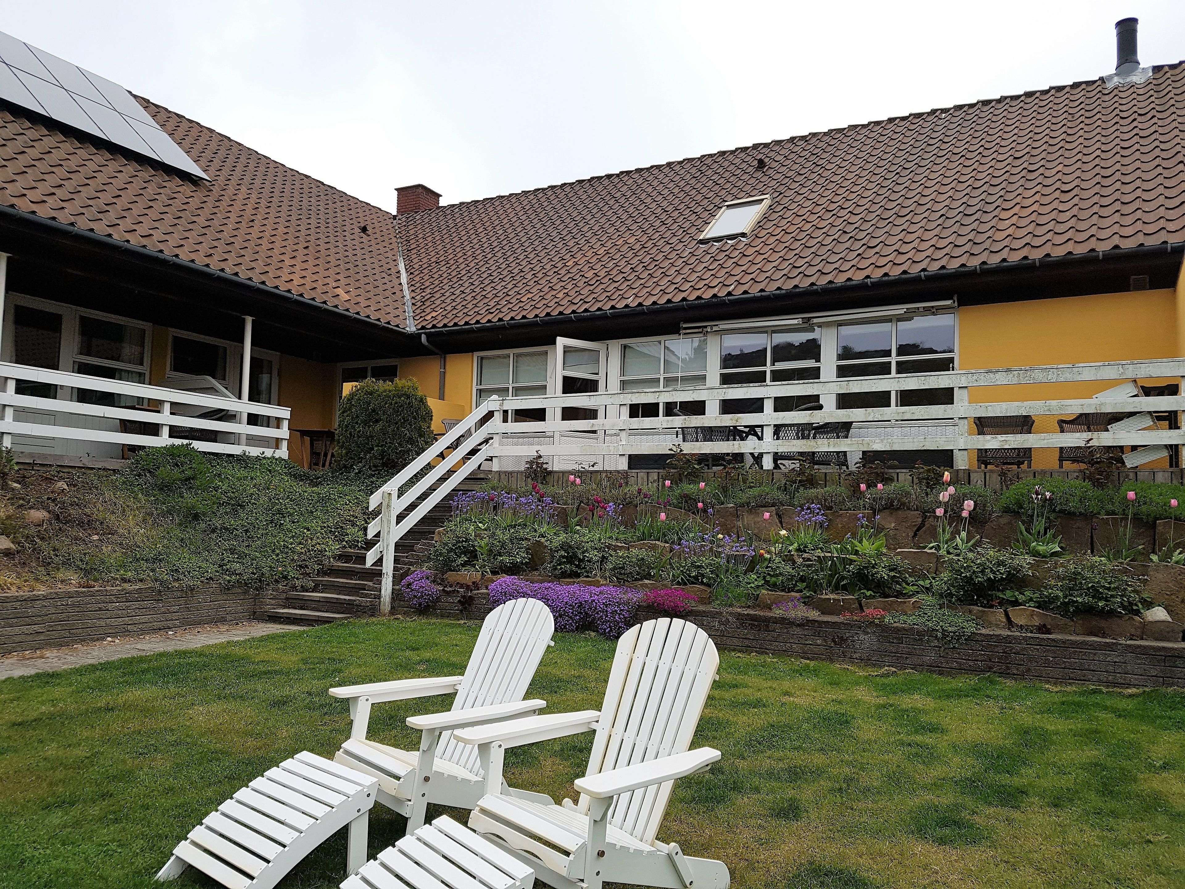 A house with a sloped roof, a front porch with white railings, and a garden with various plants and flowers.