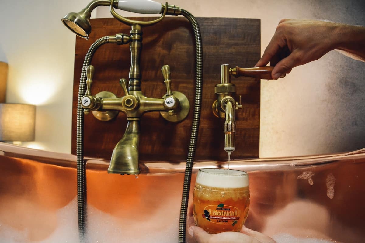 A brass faucet with a shower head and hose, a glass of beer, and a hand turning the faucet.