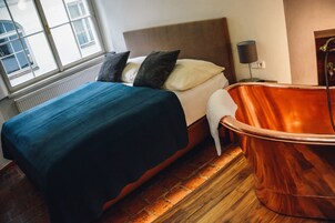 Een hotelkamer met een bed, een groot raam, een radiator, een houten vloer en een bad.