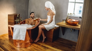 Un homme et une femme, dans un décor de spa, sont assis dans une baignoire en cuivre, avec un banc en bois et un grand récipient en cuivre posé sur un comptoir en bois.
