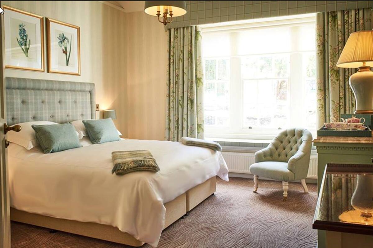 Une chambre spacieuse avec un grand lit, une chaise, une table avec une lampe et deux reproductions botaniques encadrées accrochées au mur.