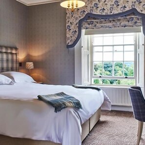 A bedroom with a bed, a window with a view of greenery, a chair, and a wall-mounted lamp. - Walwick Hall (Hexham)