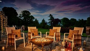 Een terras met witte stoelen en geruite kussens, een vuurkorf, kaarsen en flessen drank.