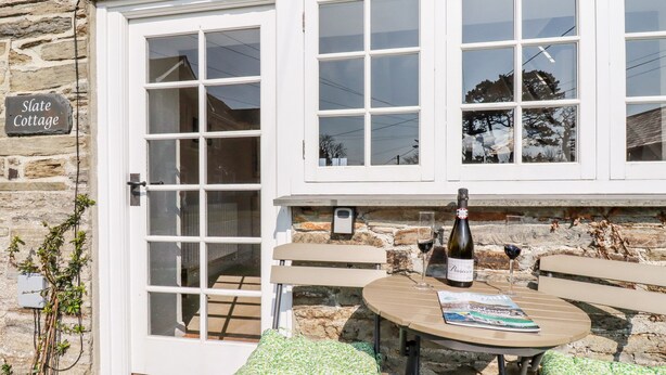 Ein Steinhäuschen mit einem Schild, auf dem "Slate Cottage" steht. Es hat eine weiße Tür mit Glasfeldern, einen kleinen Tisch mit einer Weinflasche und einem Glas sowie zwei gepolsterte Bänke.