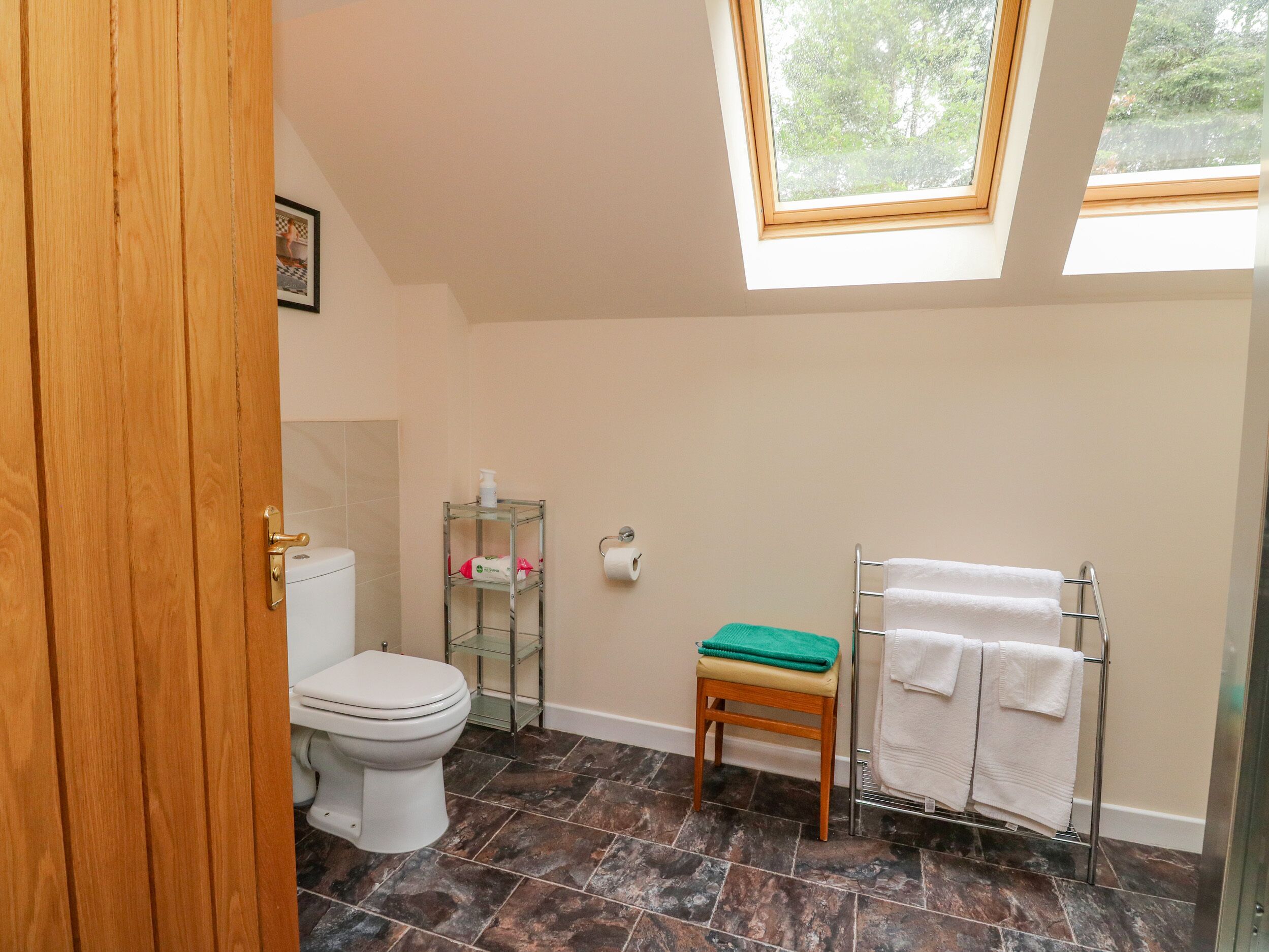 Et badeværelse med ovenlys, toilet, hylder med toiletartikler, en lille skammel og et håndklædestativ.