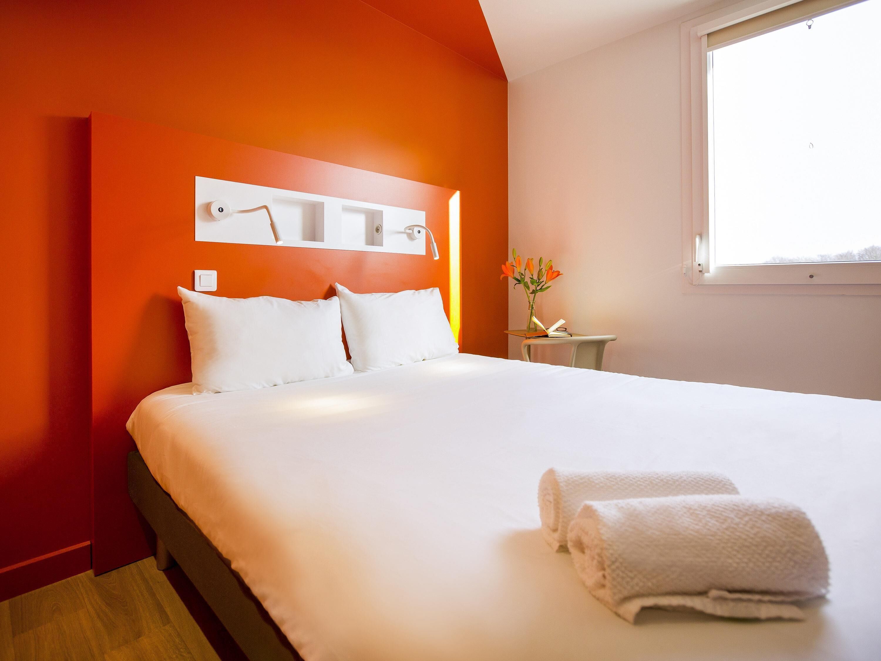 Una habitación de hotel con una cama grande, una mesita de noche con lámpara, un jarrón con flores y una ventana.