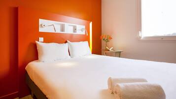Una habitación de hotel con una cama grande, una mesita de noche con lámpara, un jarrón con flores y una ventana.