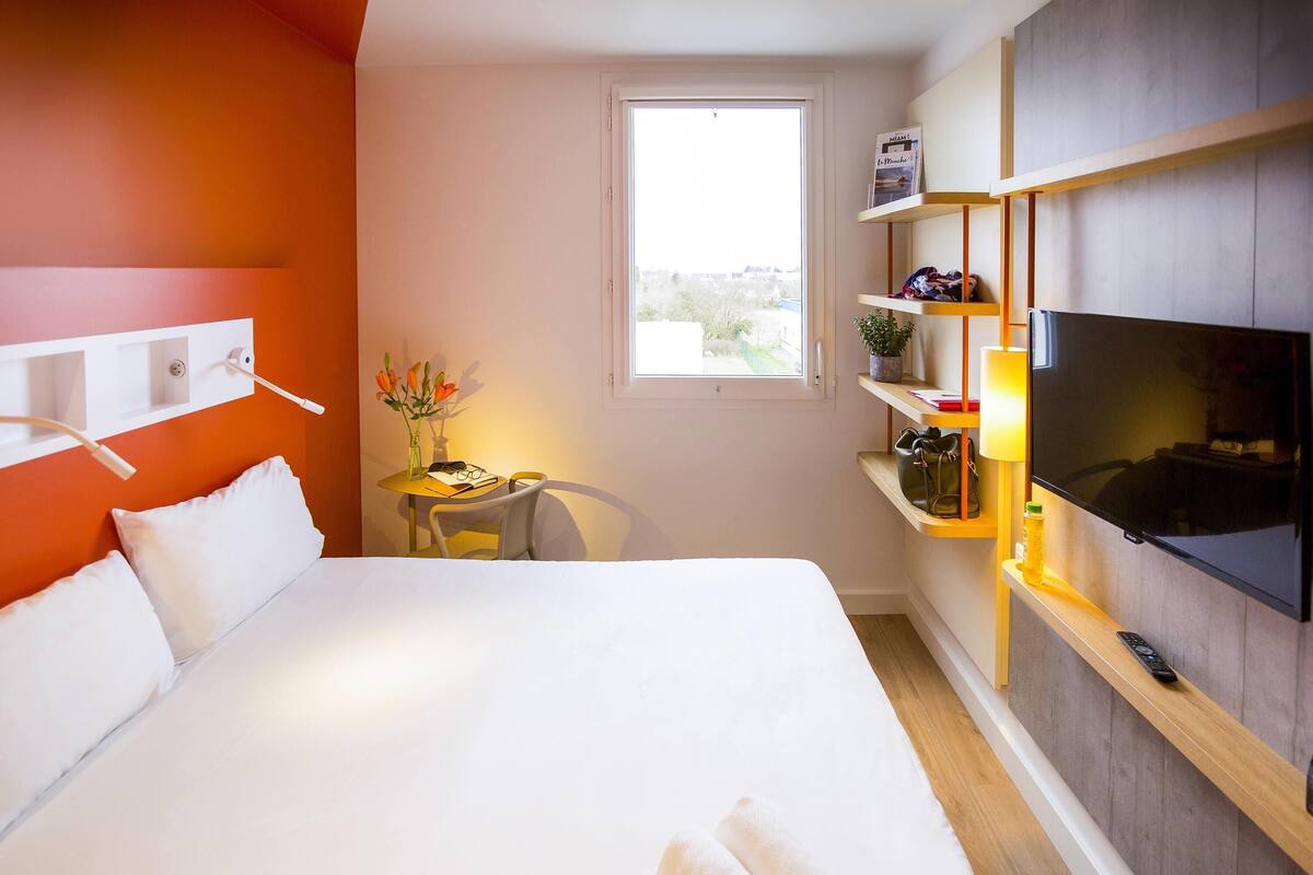 A hotel room with a large bed, a desk with a chair, a TV mounted on the wall, and a window.