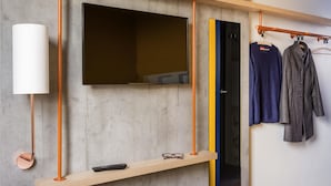 Una habitación moderna con un televisor montado, un estante de madera con un control remoto y un perchero con dos prendas.