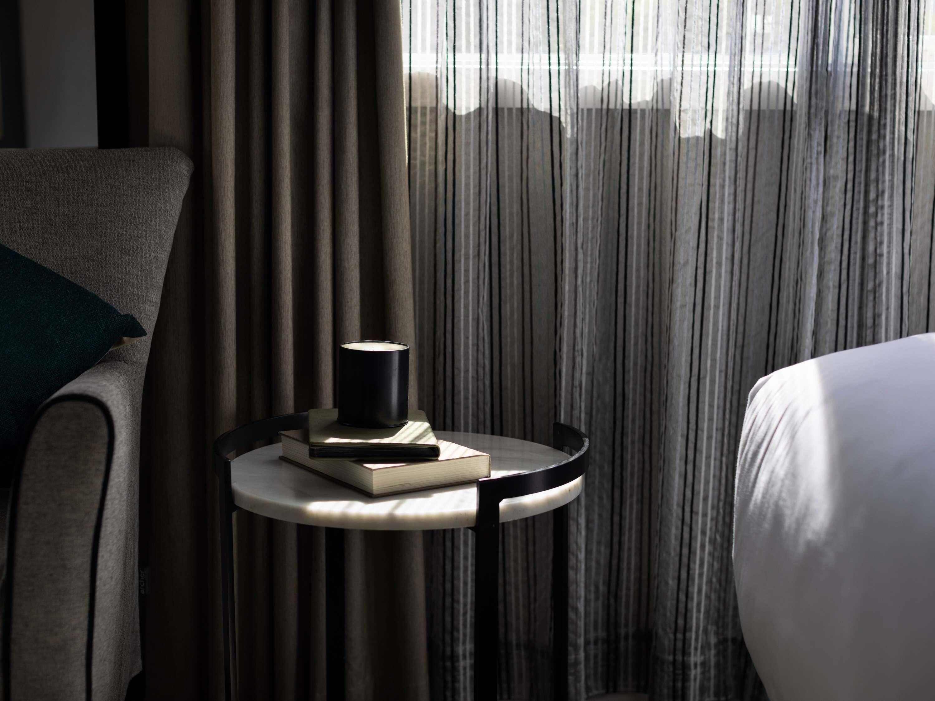 Habitación de hotel con cama, una mesa redonda con un libro y una vela, y vistas a través de cortinas translúcidas.