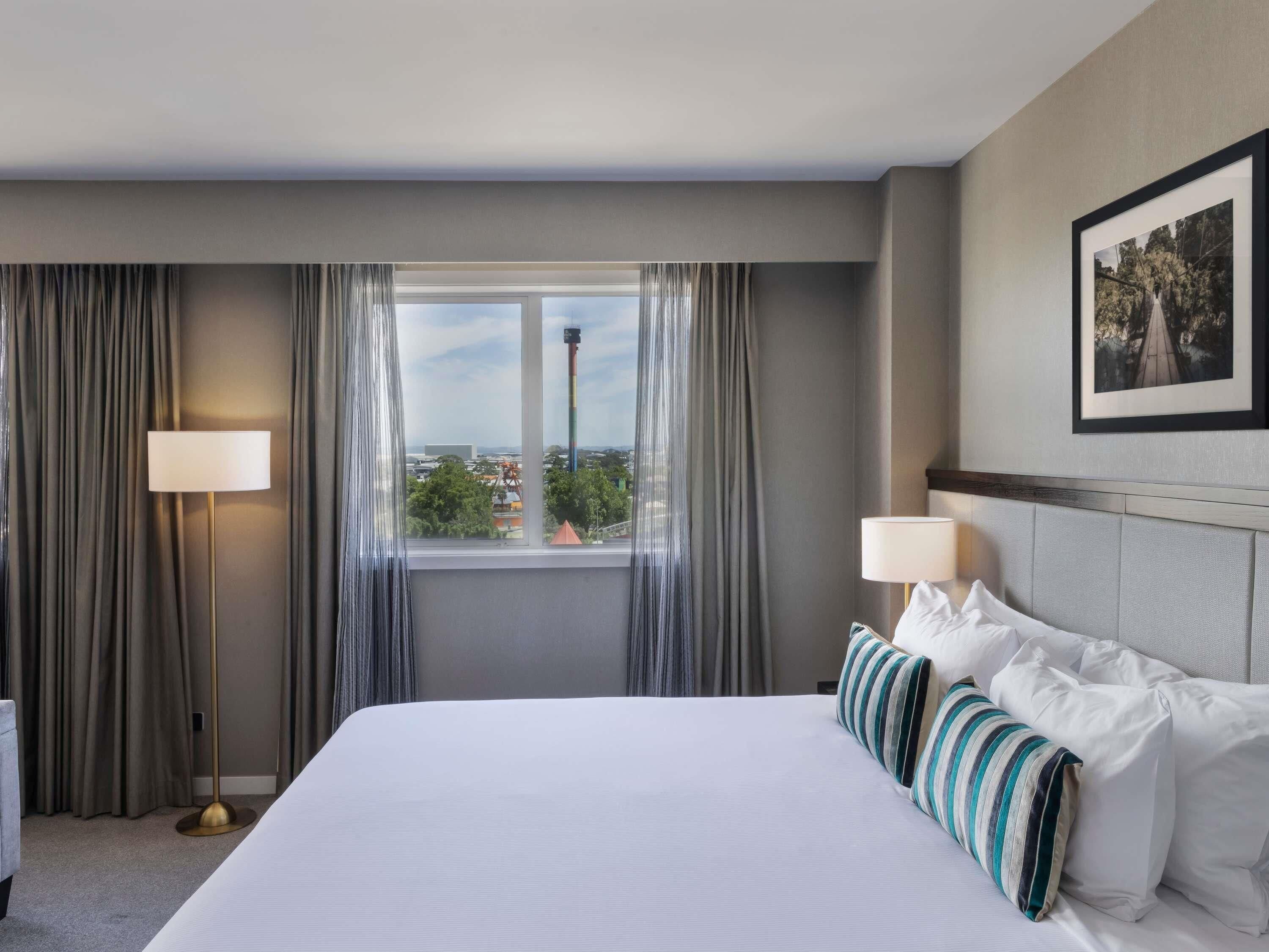 A hotel room with a large bed, two bedside lamps, a window with a view of a cityscape, and a framed picture on the wall.