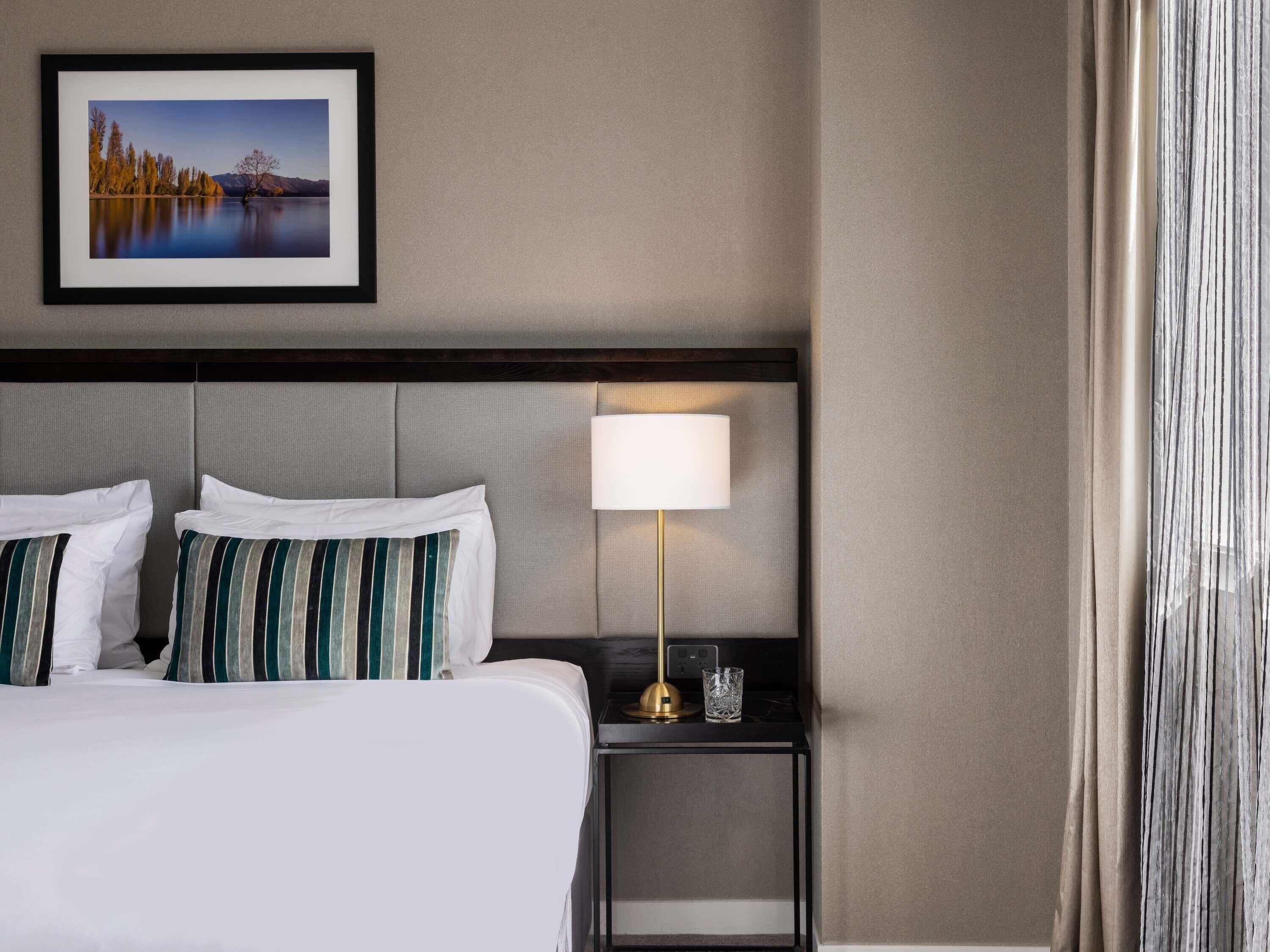 Habitación de hotel con cama, almohadas, mesita de noche con lámpara, un cuadro enmarcado en la pared y una ventana con cortinas.