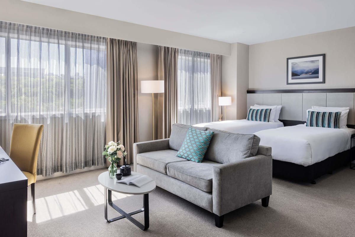 A hotel room with a sofa, two beds, a small table with a book, and a lamp.