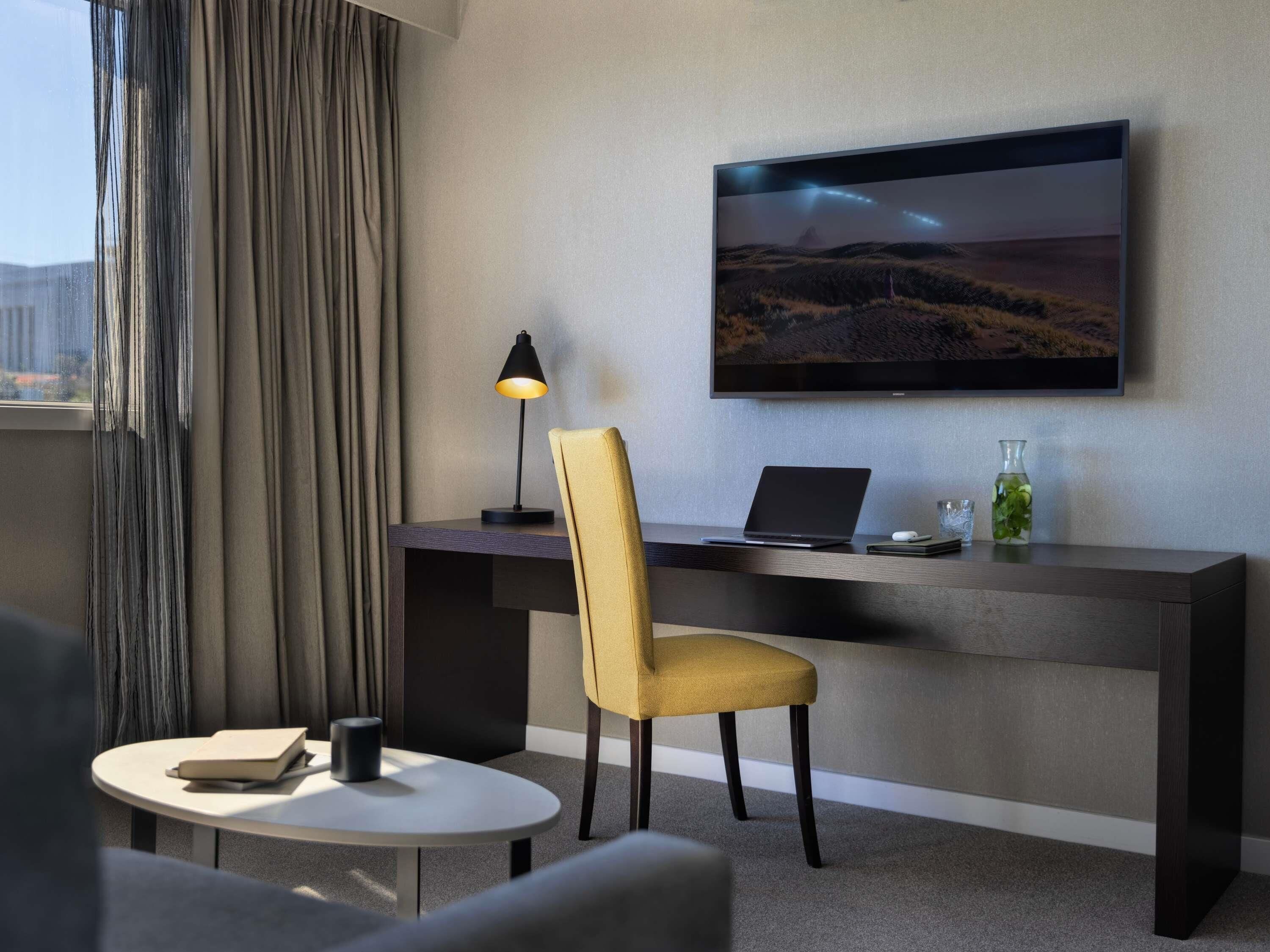 A hotel room with a desk, chair, laptop, TV, and a small table with a book and a cup.