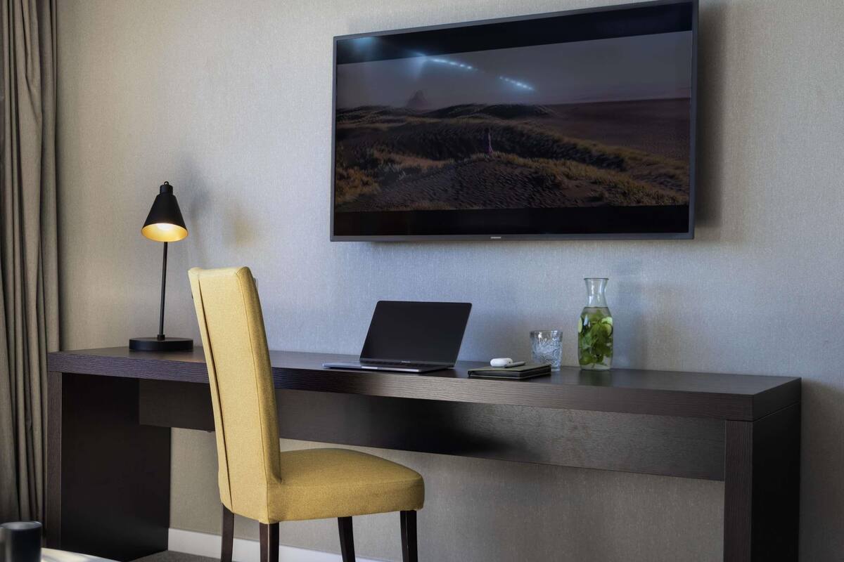 A modern hotel room with a desk, laptop, and a television displaying a landscape.