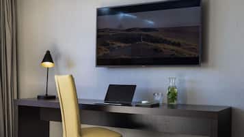 A modern hotel room with a desk, laptop, and a television displaying a landscape.