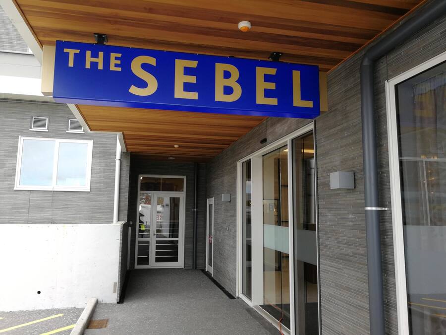 A building entrance with a sign reading "THE SEBEL" and a covered walkway leading to a glass door entrance.