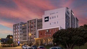 A multi-story hotel with a modern facade, a prominent sign for 'The Sebel,' and a parking area with several cars.