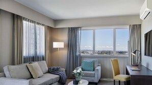 A hotel room with a sofa, armchair, a small table with a vase, a lamp, and a desk with a laptop.