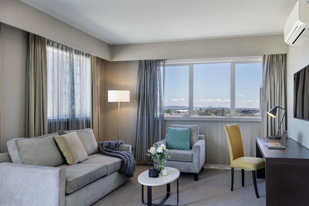 A hotel room with a sofa, armchair, a small table with a vase, a lamp, and a desk with a laptop.