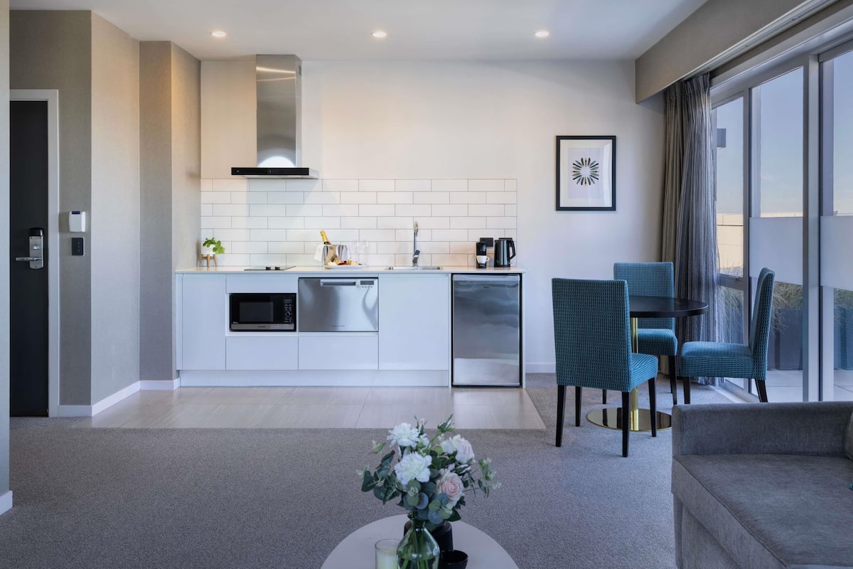 A modern kitchen with built-in appliances, a dining area with blue chairs, and a large window with a view.