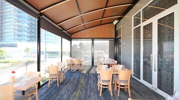 A covered outdoor dining area with tables and chairs, large windows, and a view of the cityscape.