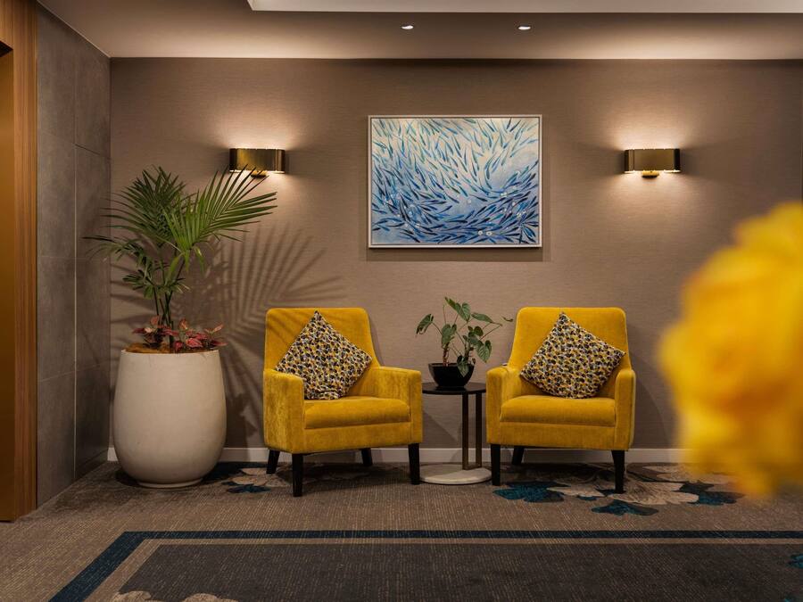 Un hall d’entrée avec deux fauteuils jaunes, une grande plante en pot, un tableau accroché au mur et deux appliques murales.