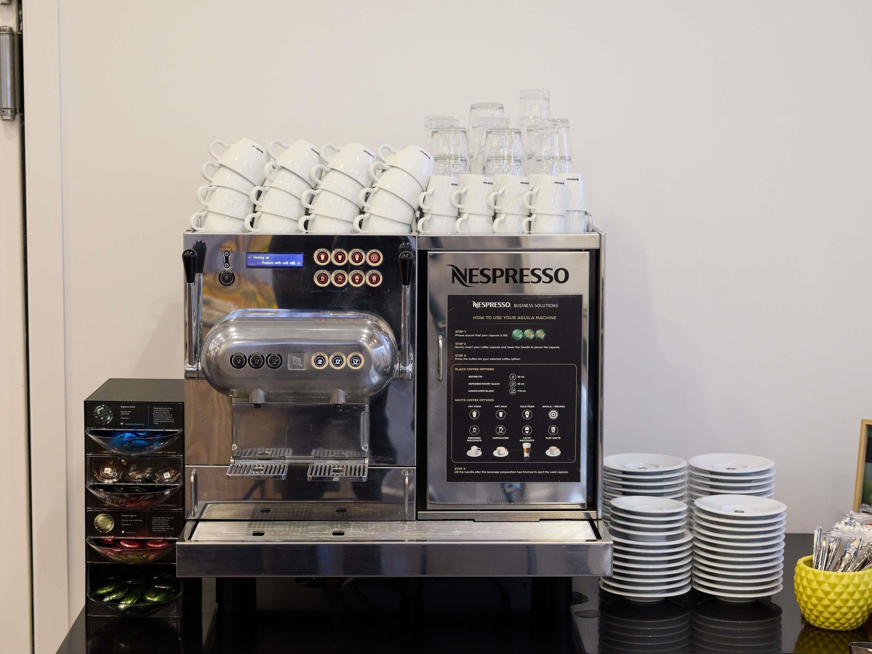 Una macchina per caffè Nespresso con diverse tazze impilate sopra, un distributore di capsule e una pila di piatti e bicchieri sul bancone.