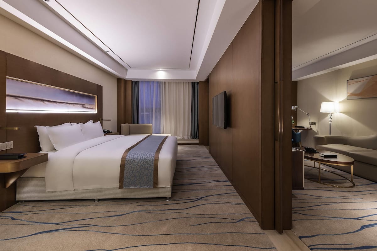 A hotel room with a large bed, a desk with a lamp, a TV, and a seating area with a couch and chair.