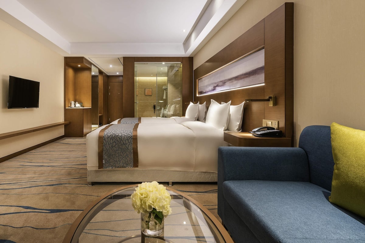 A hotel room with a bed, a sofa, a TV, a mirror, and a vase with flowers.