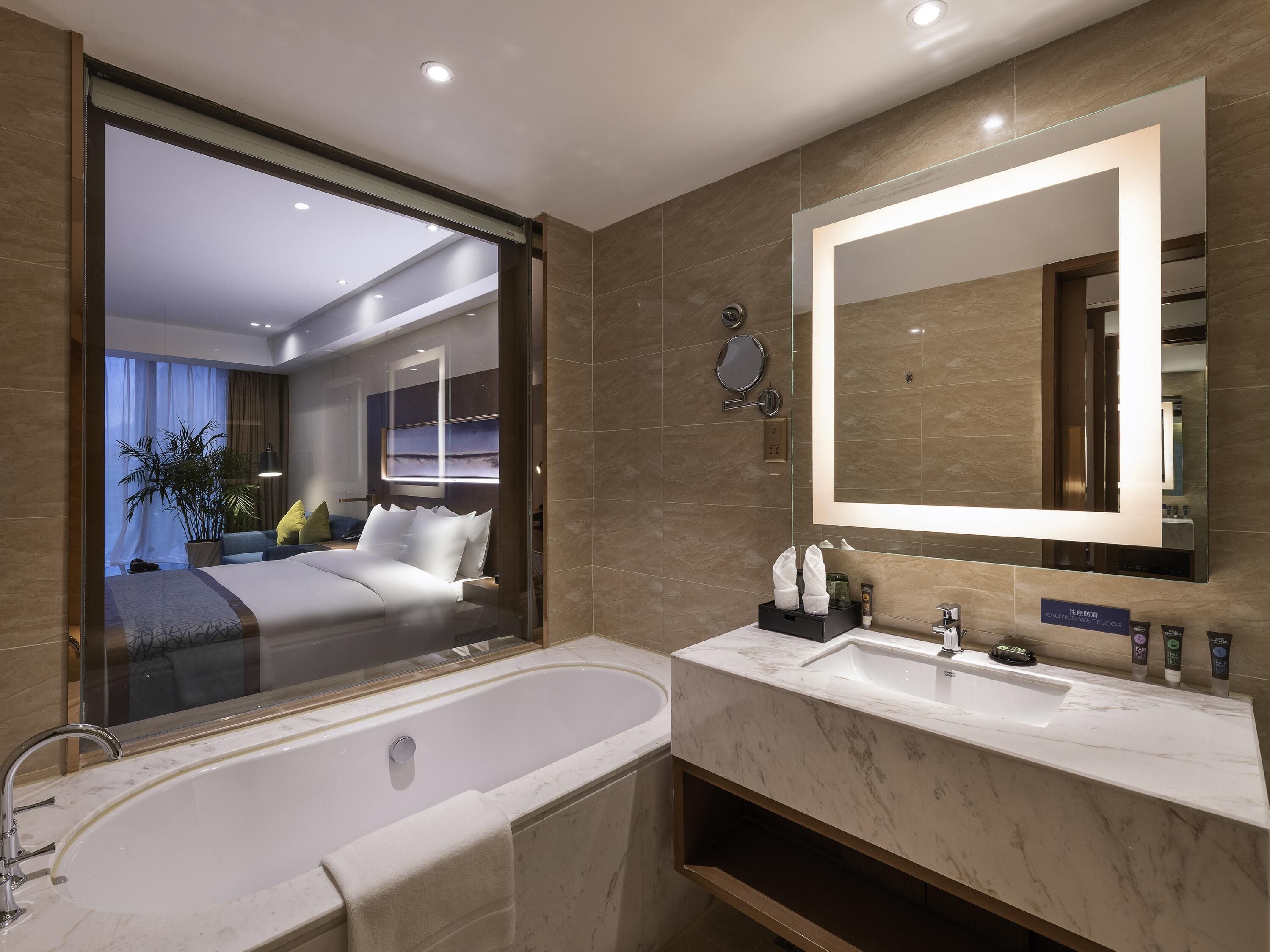 Un baño de hotel moderno con una bañera grande, un tocador con espejo y vistas del dormitorio a través de una puerta de cristal.