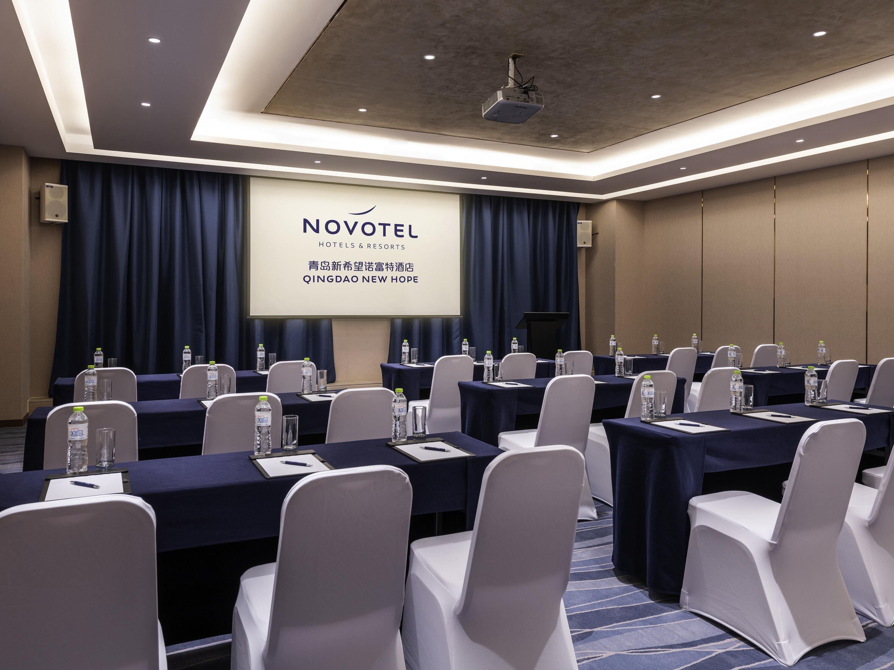 Un salón de conferencias con mesas, sillas y una pantalla proyectora mostrando 'NOVOTEL HOTELS & RESORTS' y 'QINGDAO NEW HOPE'.