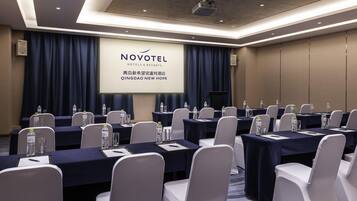 Un salón de conferencias con mesas, sillas y una pantalla proyectora mostrando 'NOVOTEL HOTELS & RESORTS' y 'QINGDAO NEW HOPE'.