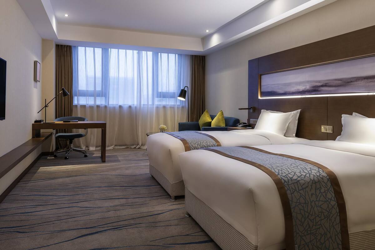 A hotel room with two beds, a desk, a chair, a lamp, and a large window with curtains.