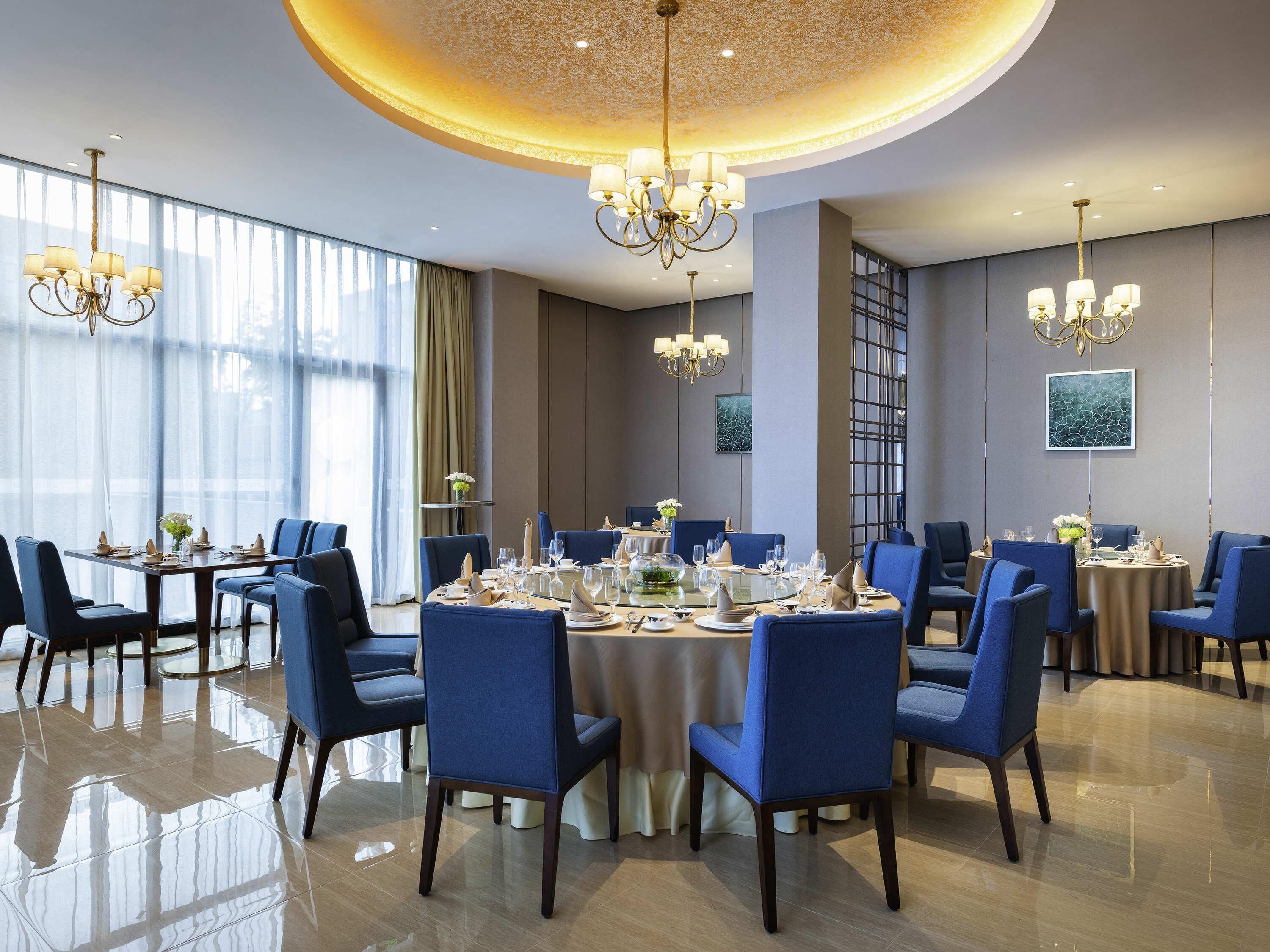 A dining area with round tables, blue upholstered chairs, chandeliers, and a large window with sheer curtains.