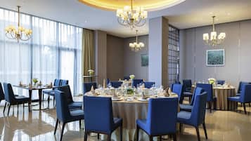 A dining area with round tables, blue upholstered chairs, chandeliers, and a large window with sheer curtains.