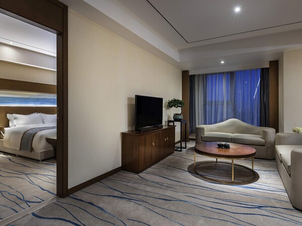 A modern hotel room with a bed, a television, a sofa, a coffee table, and a large window with curtains.
