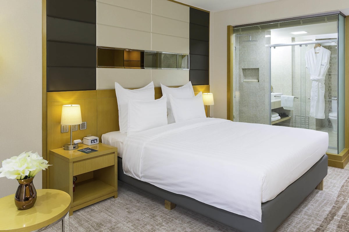 Habitación de hotel con una cama grande, lámparas de noche, una mesita de noche, un jarrón con flores y zona de ducha con paredes de vidrio.