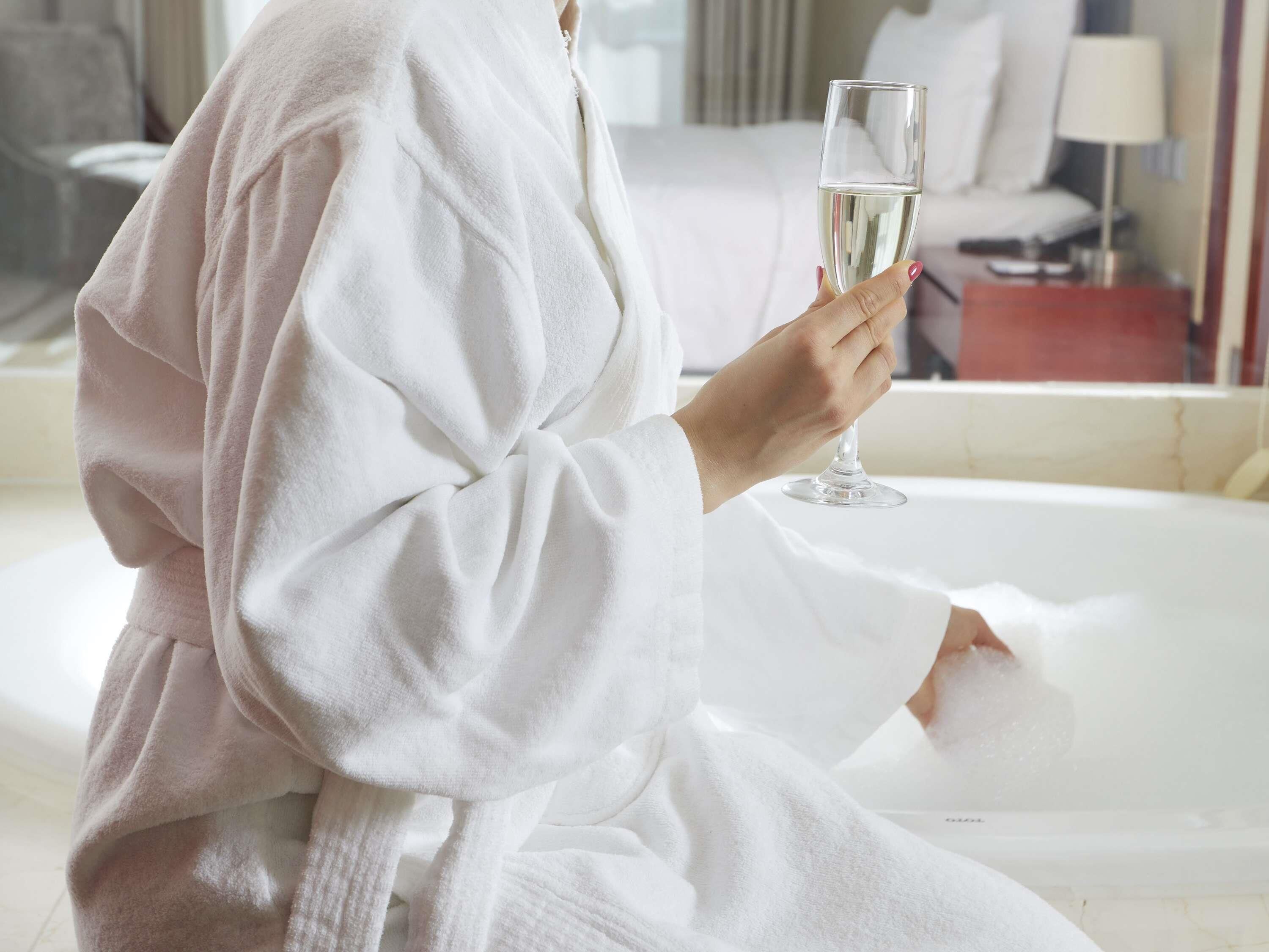 Une personne vêtue d’un peignoir blanc est assise dans une baignoire, un verre de champagne à la main.